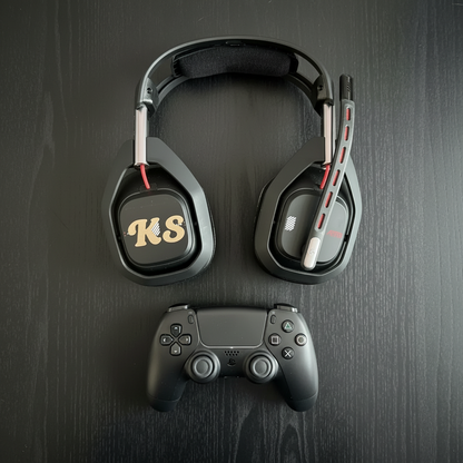 A black over-ear gaming headset with a flexible microphone and red accents, shown on a dark wood surface with a game controller partially visible at the bottom. The earcups feature a gold 'KS' logo.